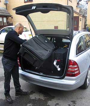 slight angled photo of a airport taxis driver loading a customers luggage into a silver coloured mercedes c class estate car cab just before setting off to manchester airport